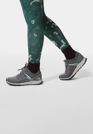 Grijze sportschoenen met groene accenten, voorzien van een gemêleerde bovenkant, een gevoerde zool en duurzame grip. Gepaard met donkere, kleurrijke leggings.