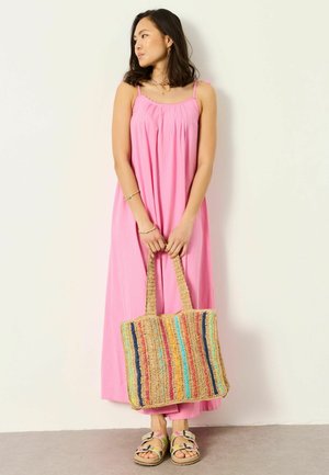 Woman in a pink sleeveless maxi dress holding a multicolored woven tote bag, wearing floral sandals, standing against a plain wall.