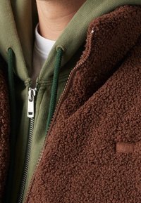 Ruskea fleecetakki, jossa on teksturoitu pinta, vetoketjulla suljettava etuosa ja vihreä huppari alla, jonka alla on valkoinen paita.