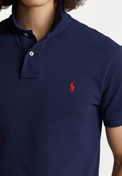 Tums zila polo krekls no teksturēta kokvilnas, ar divu pogu aizdari un mazu sarkanu izšūtu logotipu uz kreisā krūts. Apkakle ir pacelta.