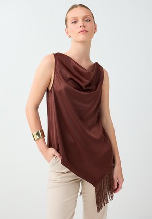 Femme portant un haut en satin marron sans manches avec un ourlet asymétrique à franges, un pantalon beige, un bracelet manchette doré et de petites boucles d'oreilles en anneau.