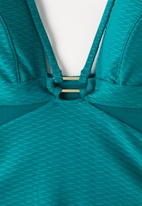 Turkoza sporta krekls ar teksturētu rakstu, kam ir regulējamas siksnas un zelta rāvējslēdzēja detaļa aizmugurē. Gluda auduma apdare.