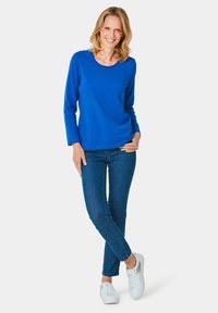 Blaues Langarm-Oberteil mit runder Ausschnitt, kombiniert mit engen blauen Jeans und weißen Turnschuhen, zeigt ein lässiges Outfit.