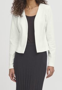 Veste blanche courte en tissu lisse, avec un col rond et des manches longues, portée sur un haut sombre et une jupe plissée.