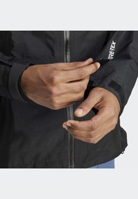 Giacca nera impermeabile con zip frontale, polsini in Velcro e logo decorativo. Il tessuto sembra texturizzato e durevole.