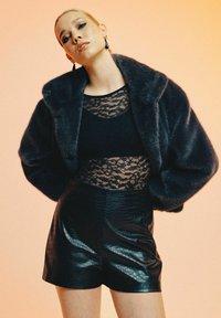 Veste en fausse fourrure marine sur un haut en dentelle noire, associée à des shorts en simili-crocodile noirs. Des textures et des motifs distincts sont visibles.