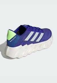 Sneaker viola con tomaia in rete testurizzata, logo a tre strisce bianche e un accento verde a contrasto sul tallone. Presenta una suola bianca spessa.