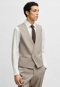 Gilet beige con un motivo testurizzato, due bottoni e tasche laterali, indossato sopra una camicia bianca e una cravatta marrone scuro. Vestibilità su misura.