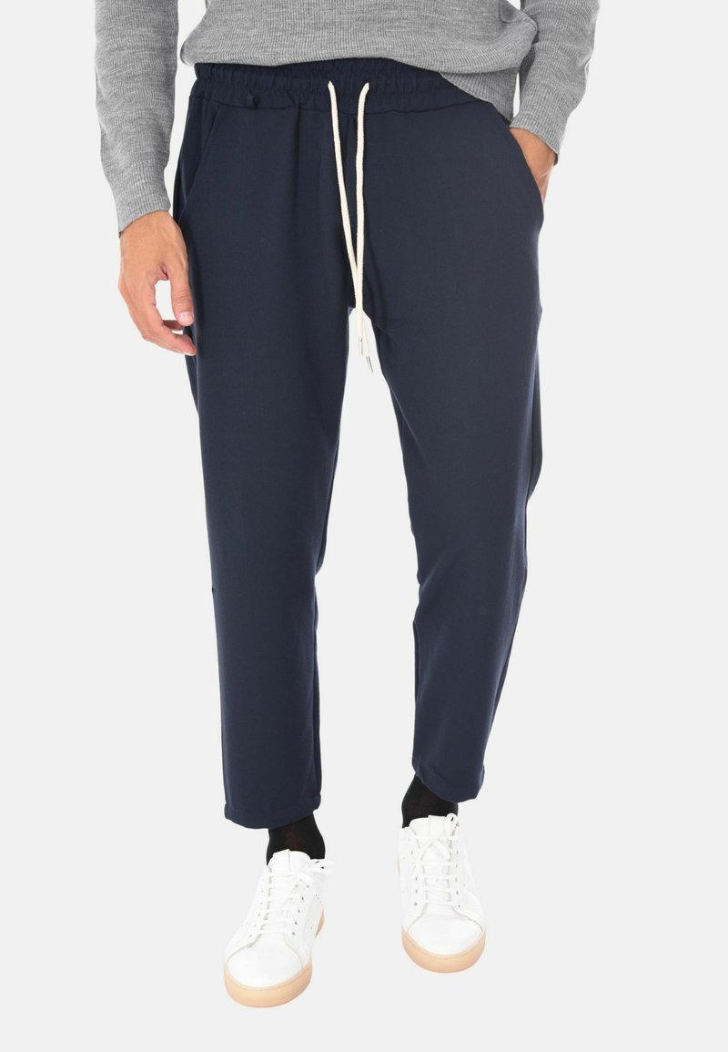 Uomo che indossa pantaloni blu navy con cordoncino, maglione grigio, calze nere e sneakers bianche, in piedi con una mano in tasca.