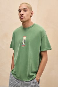 Young man with short blond hair wears green graphic t-shirt and black-and-white checkered pants, hands in pockets, neutral beige background.