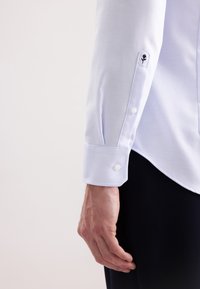 Camicia azzurra chiara con motivo a spina di pesce, polsini con bottoni e dettaglio floreale ricamato sul polsino. Tessuto dalla texture liscia.