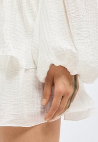 Χέρι που ακουμπά στον μηρό, φοράει χαλαρό λευκό υφασμάτινο ύφασμα με μαζεμένο μανικοκόφτη και κοντή γραμμή.