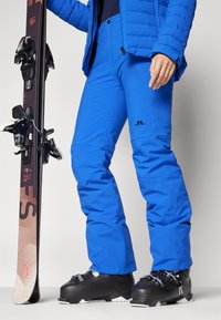 Μπλε σκι παντελόνι με λεία υφή, διαθέτει λογότυπο, κλείσιμο με κουμπί και ανατομικά γόνατα. Συνδυάζεται με μαύρες μπότες σκι και σκι.