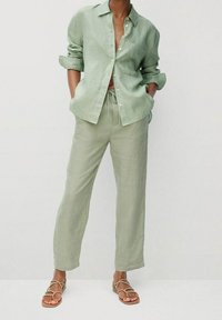 Jasnozielona lniana koszula z kieszenią i podwiniętymi rękawami, zestawiona z dopasowanymi lnianymi spodniami i brązowymi sandałami na ramiączkach. Luźny krój i faktura.