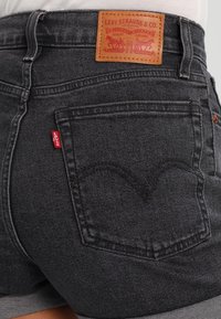 Bermudas de mezclilla gris oscuro con un parche trasero de piel que presenta el logotipo de la marca, una etiqueta roja en el costado y un patrón de costura distintivo en los bolsillos.