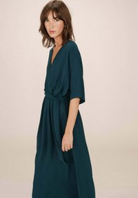 Teal wikkeljurk van een lichtgewicht stof, met korte mouwen, een diepe V-hals en een strikdetail bij de taille, die tot aan de enkels valt.