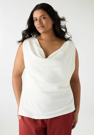 Donna con capelli lunghi e scuri che indossa una maglia bianca senza maniche drappeggiata e pantaloni color ruggine, su uno sfondo chiaro e uniforme.