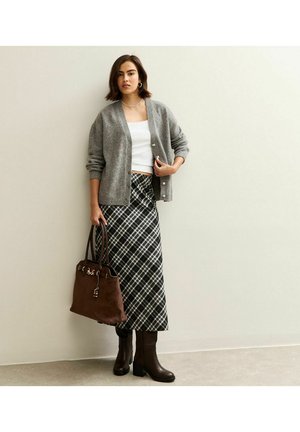 Cardigan en tricot gris avec des boutons sur un crop top blanc, associé à une jupe maxi à carreaux et des bottes à talons marron. Sac à main marron avec des accents.