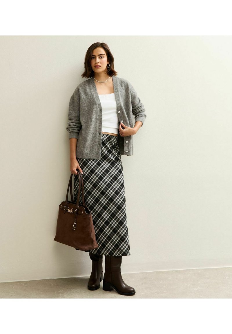 Cardigan de punto gris con botones sobre un top corto blanco, combinado con una falda maxi de cuadros y botas de tacón marrones. Bolso marrón con detalles.