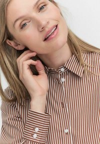 Chemise à rayures brunes et blanches avec un col pointu et des poignets à boutons, portée par une personne ayant les cheveux longs.