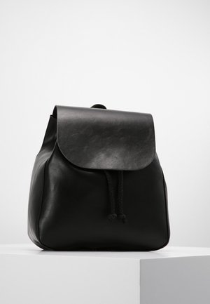 Black leather backpack with a flap cover and drawstring closure, placed on a white pedestal against a plain light background.