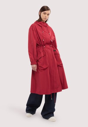 Frau trägt einen langen roten Kapuzenmantel mit großen Fronttaschen, kombiniert mit weit geschnittenen dunklen Hosen und weißen Turnschuhen, steht vor einem schlichten Hintergrund.