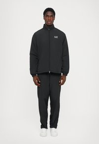 Black zip-up jacket with a high collar, featuring a logo on the left chest. Paired with matching black pants and white sneakers.