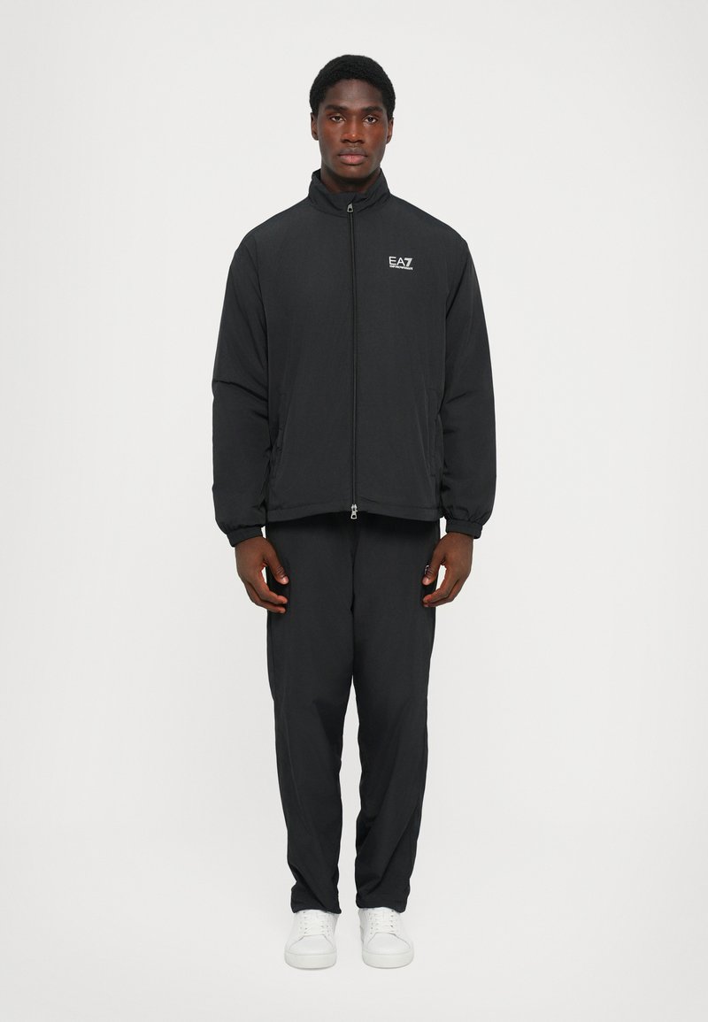 Black zip-up jacket with a high collar, featuring a logo on the left chest. Paired with matching black pants and white sneakers.