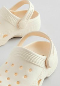 Zuecos de color beige claro, fabricados en un material flexible, con orificios de ventilación, una banda texturizada y una correa trasera ajustable para un mejor ajuste.