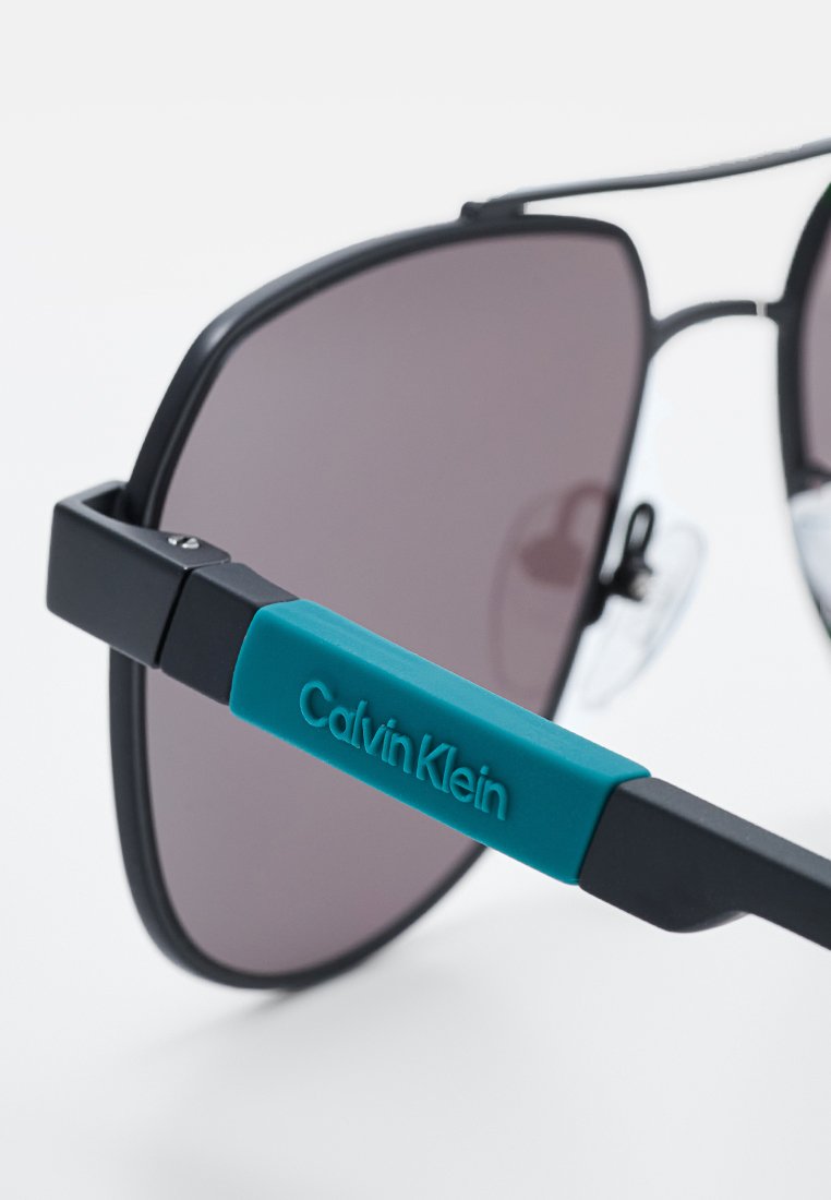 Gafas de sol de estilo black metal con grandes lentes grises. Presentan un brazo de plástico teal etiquetado como "Calvin Klein". Textura suave y diseño geométrico.