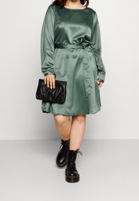 Abito in satin verde con maniche lunghe e vita regolabile, abbinato a una pochette nera e stivali neri con lacci, che mostrano una texture liscia.