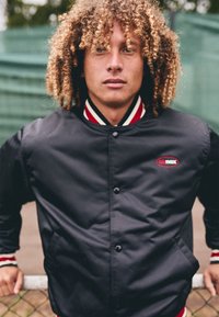 Black satin jacket with red and white striped accents on the collar and cuffs. Features a logo on the left chest and button closure.