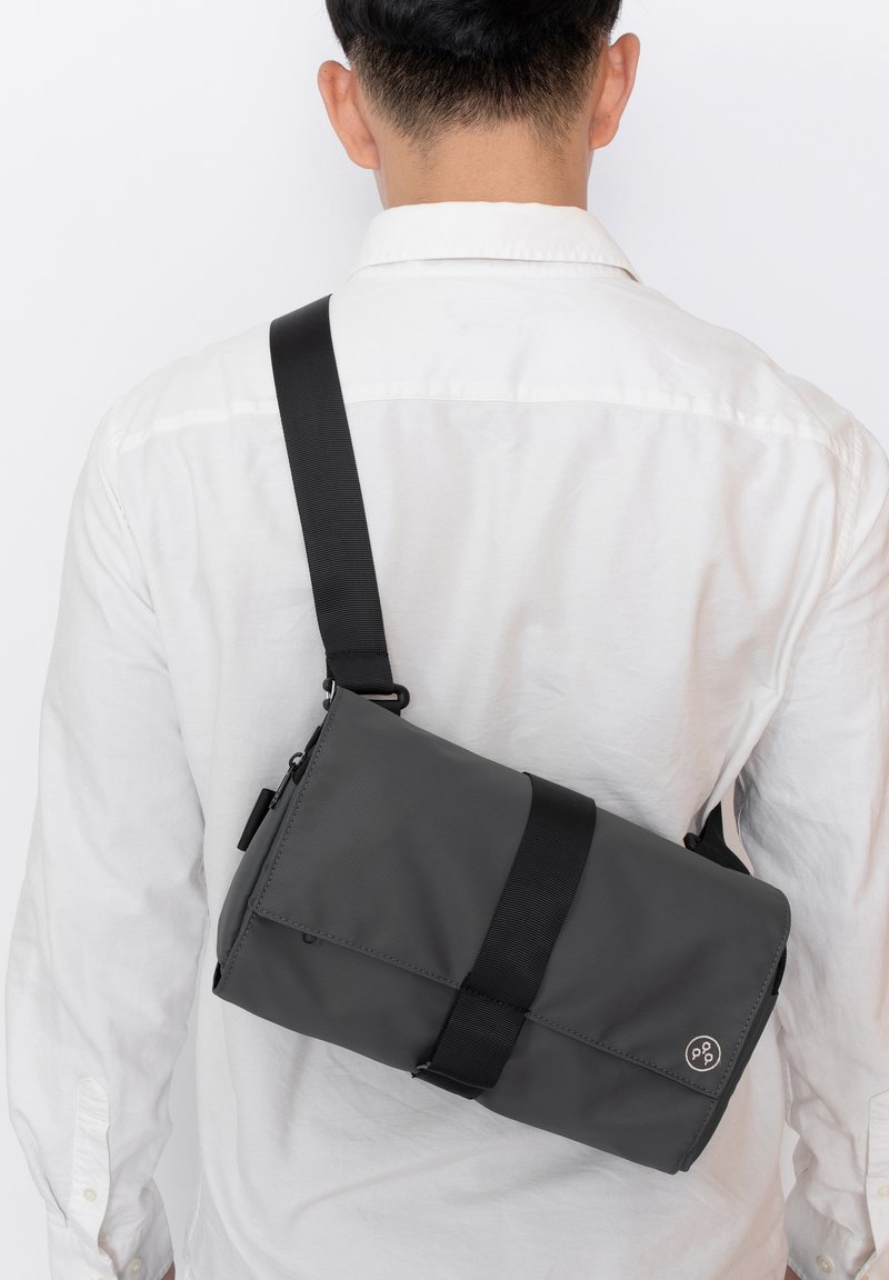 Borsa a tracolla grigia realizzata in tessuto resistente, dotata di una larga tracolla, chiusura a patta e un accento con logo circolare.