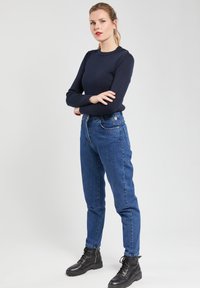 Pull à col rond bleu marine à manches longues associé à un jean denim bleu taille haute et des bottes à lacets noires. Présente des boutons à l'épaule.