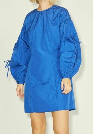 Mujer con un vestido azul brillante con mangas largas abullonadas atadas con lazos y un escote redondo sencillo.