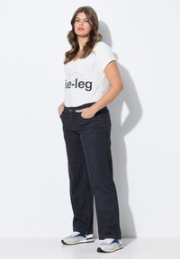Witte V-hals t-shirt met zwarte tekst, gecombineerd met donkerblauwe wijde jeans. Het model draagt stijlvolle sportieve sneakers met meerkleurige accenten.