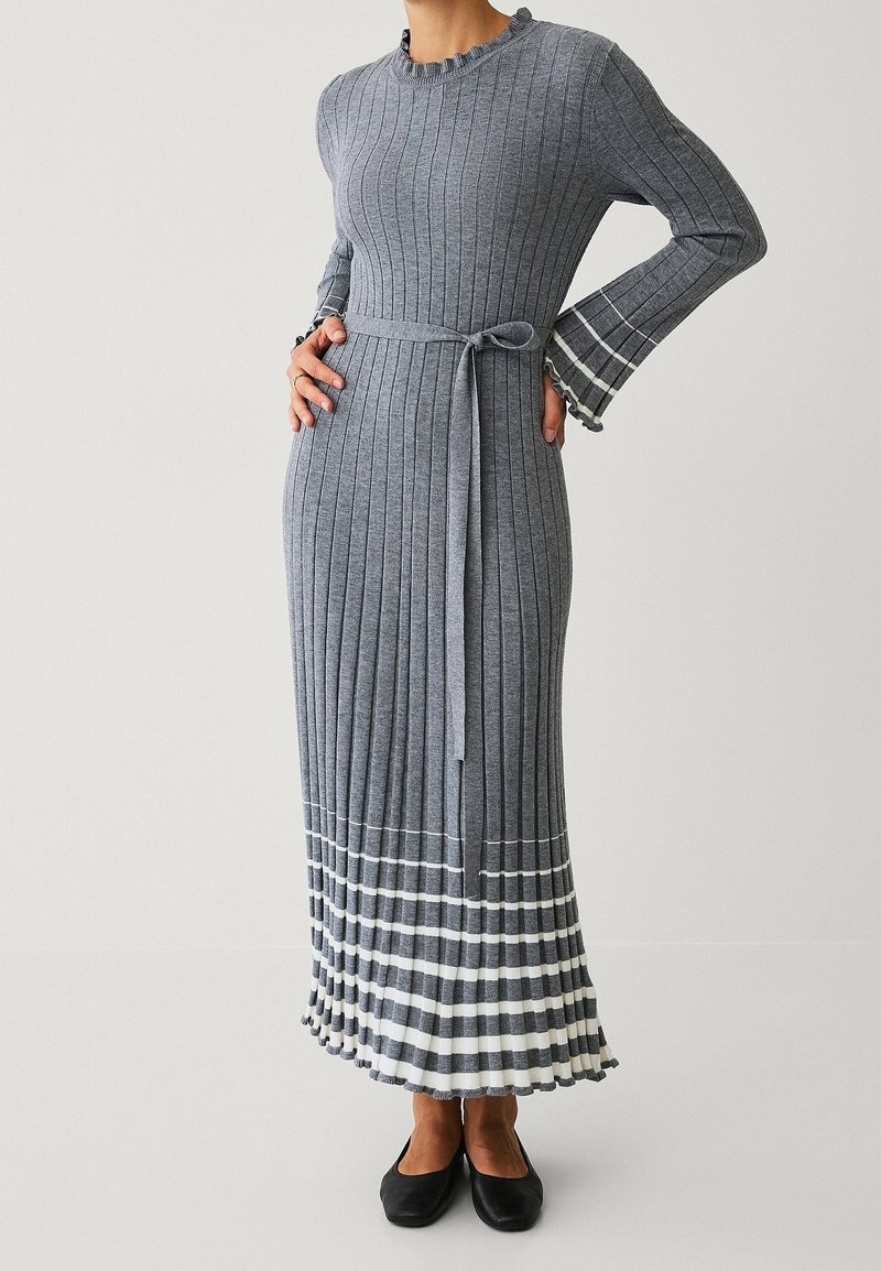 Robe en maille côtelée grise à manches longues avec des rayures blanches à l'ourlet et aux poignets, ceinture nouée à la taille, portée avec des chaussures plates noires.