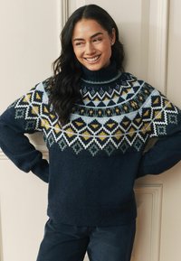 Tumši zils adīts džemperis ar augstu apkakli, ar ģeometriskiem rakstiem zaļā, baltā un dzeltenā krāsā krūšu un piedurkņu rajonā.