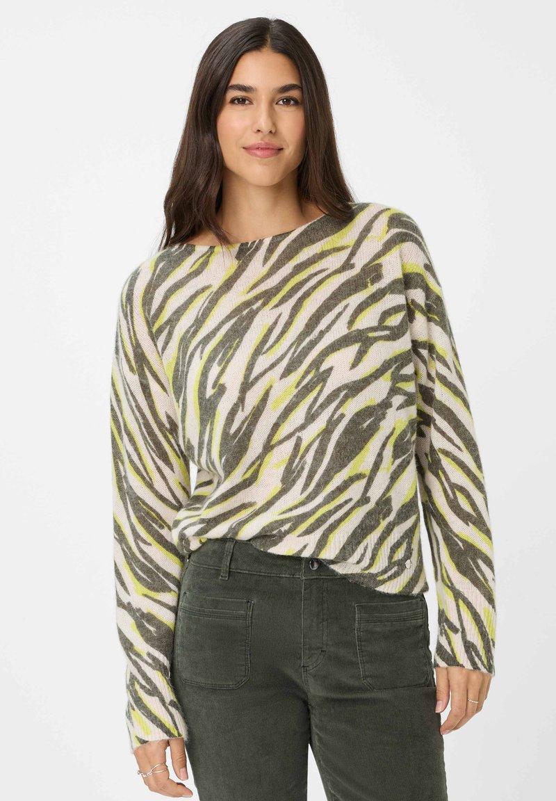 Frau mit langen dunklen Haaren, die einen lockeren zebra-gemusterten Pullover in Grün- und Grautönen sowie eine dunkelgrüne Cordhose trägt.