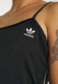 Črni športni top s tankimi naramnicami. Ima belo izvezeno logo na levi prsni strani in stranske detajle. Gladek, raztegljiv material.