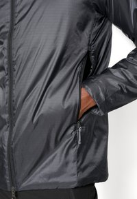 Schwarze leichte Jacke aus glattem, glänzendem Stoff. Verfügt über Reißverschlusstaschen und ein strukturiertes Design. Enganliegend mit langen Ärmeln.
