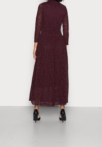 Burgundy spetsklänning med långa ärmar och vid kjol, prydd med intrikata blommönster, bärs med svarta högklackade skor.