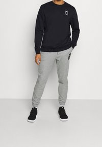 Czarna bluzka z kapturem z logo na lewej piersi, w połączeniu z szarymi zwężającymi się spodniami dresowymi i czarnymi butami sportowymi z białymi podeszwami.