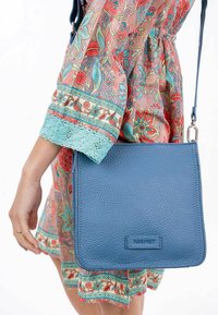 Borsa a tracolla in pelle blu testurizzata con design piatto e lunga tracolla, presentata contro un vestito colorato con un motivo e dettagli in pizzo.