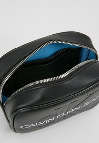 Czarna skórzana torba z wytłoczonym logo, srebrnym zamkiem błyskawicznym i jasnoniebieską podszewką. Posiada gładką fakturę i prostokątny kształt.