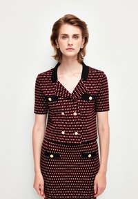 Veste à double boutonnage rouge et blanche à motifs, avec des manches courtes et un col noir. Des accents de boutons dorés et des détails de poches noires complètent le design.