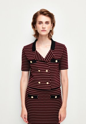 Veste à double boutonnage rouge et blanche à motifs, avec des manches courtes et un col noir. Des accents de boutons dorés et des détails de poches noires complètent le design.