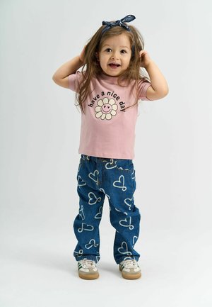 Lyserød t-shirt med teksten "hav en god dag" og et smilende blomstertryk; blå jeans med hvide hjerte mønstre; hvide sneakers med grønne striber.