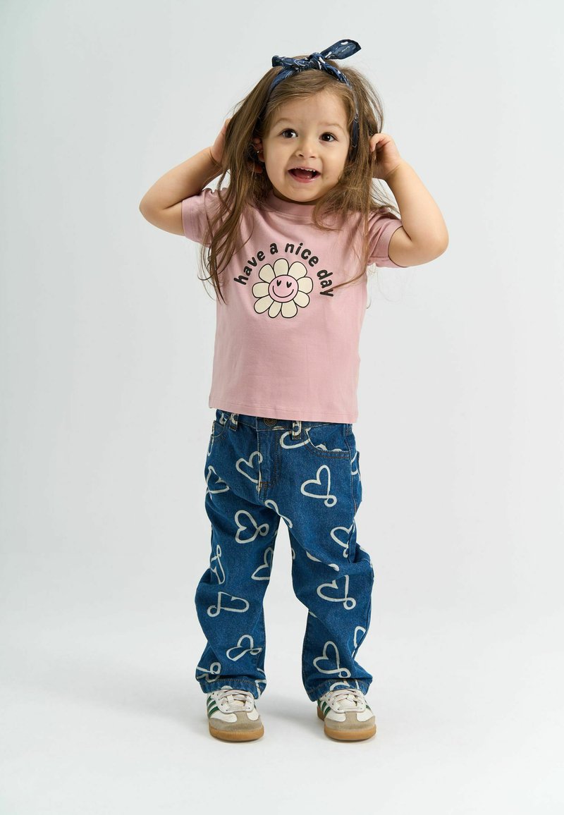 Lyserød t-shirt med teksten "hav en god dag" og et smilende blomstertryk; blå jeans med hvide hjerte mønstre; hvide sneakers med grønne striber.