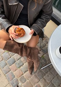 Botas marrones de ante hasta la rodilla con punta afilada, combinadas con un vestido negro y un plato de plata que sostiene un pastel de canela.